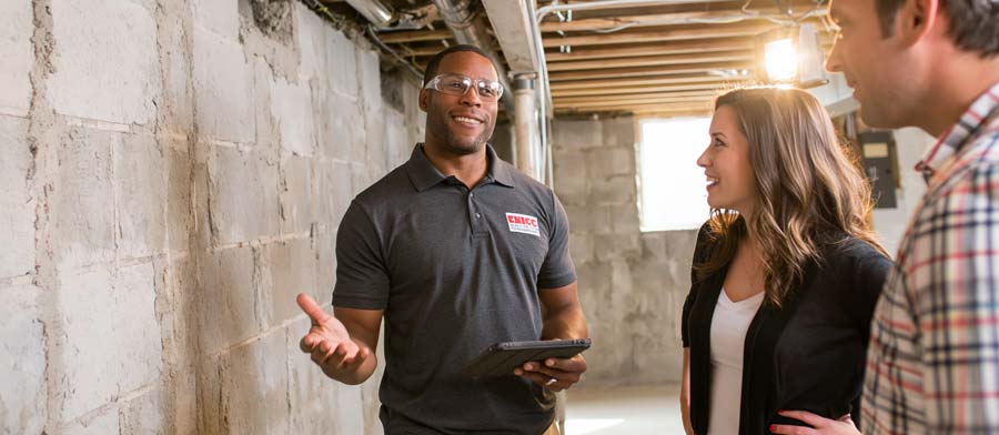 insulation contractor in company polo shirt and safety glasses holding a tablet, walking a homeowner couple in their thirties through their unfinished basement