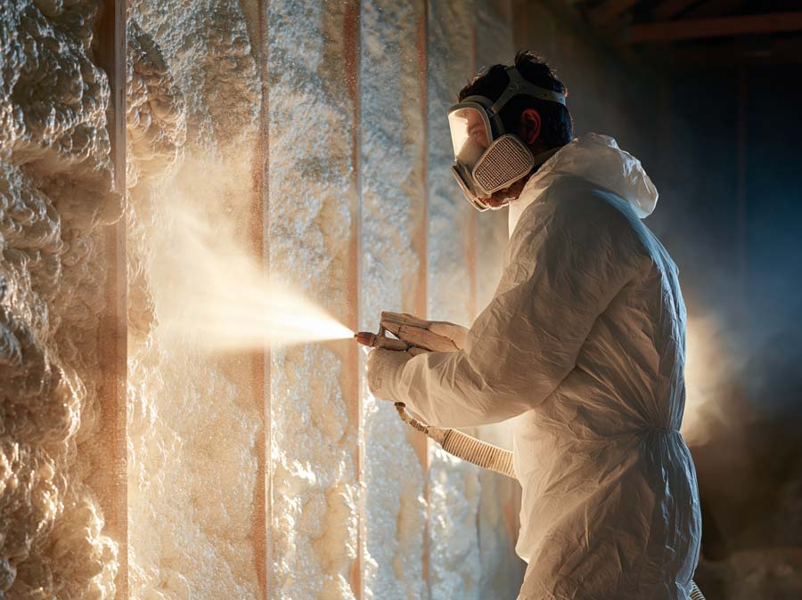 dramatic backlit shot of an insulation professional in a plain white protective suit and full-face respirator spraying foam onto an interior wall